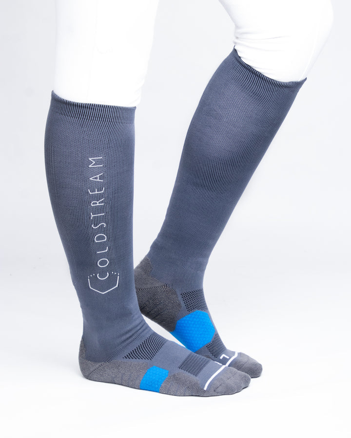A pair of knee-high athletic socks worn on a model's legs, shown against a clean white background. The socks are slate blue with gray and bright blue accents on the toes and heels, and the brand name 'COLDSTREAM' is printed vertically in white on the left leg.
