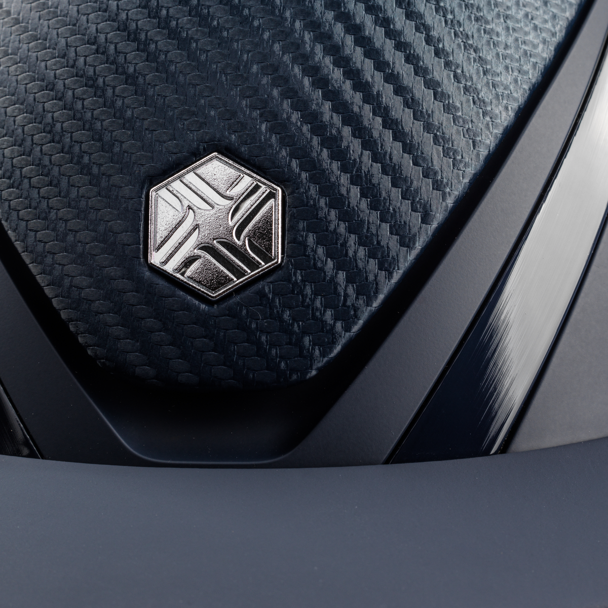 Charles Owen Hyda Carbon riding hat in Navy Matte/Navy Gloss, close-up of the brushed metal crown badge and peak